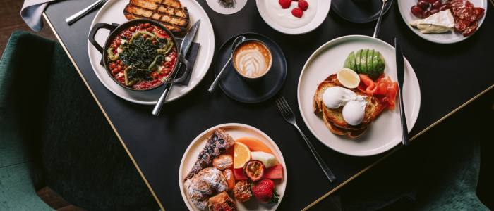 MACq01 Eat and Drink 3 2025 Table set with a variety of breakfast dishes including shakshuka with toast, poached eggs with avocado and smoked salmon, assorted pastries with fruit, a bowl of raspberries, a cheese and cured meat platter, and a cup of coffee with latte art.