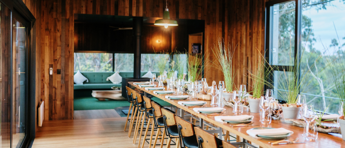 Long dining table set for a meal in a room with wooden walls and large windows. The table is decorated with plates, glasses, cutlery, and small plants, surrounded by chairs. In the background, another room with seating and large windows offers a view of trees outside.