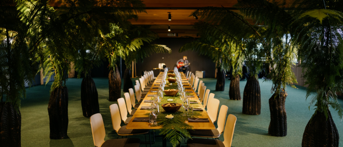 Long dining table set for a meal, surrounded by lush green ferns and plants. The table features plates, glasses, cutlery, candles, and small decorative plants. The indoor space has a green carpeted floor, wooden ceiling, and a person playing guitar in the background.
