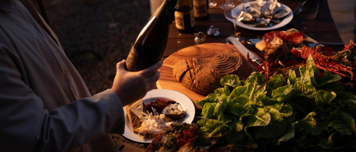 Close-up of an outdoor dining table set with seafood dishes including oysters and possibly lobster or crab, fresh greens, and a loaf of bread on a wooden cutting board. A person is holding a bottle, and the scene is lit with warm lighting, suggesting an evening gathering.