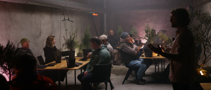 Dimly lit room with people seated at tables decorated with potted plants. A person stands at the front, appearing to speak or present. The space features bare concrete walls, a visible ceiling duct, and additional plants placed throughout