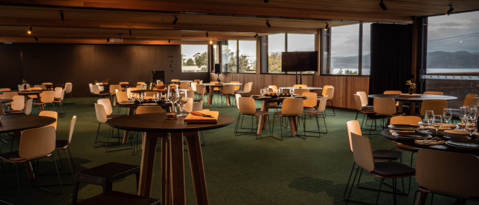 Modern restaurant dining area with round tables set with plates, glasses, and napkins. The space features wooden ceiling and wall elements, large windows providing natural light, and a clean, organized layout suitable for dining or events.