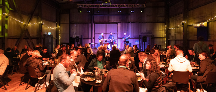 Indoor event with people seated at round tables enjoying food and drinks, while three musicians perform on a stage in the background. The venue features string lights on the walls, creating a warm, festive atmosphere.