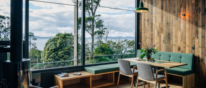 A cozy dining area with wooden walls and floors, featuring a wooden table surrounded by green upholstered bench seating and two grey chairs. Large floor-to-ceiling windows offer a scenic view of lush greenery and a distant body of water. A pendant light hangs above the table, and a small side table with books sits near the window.