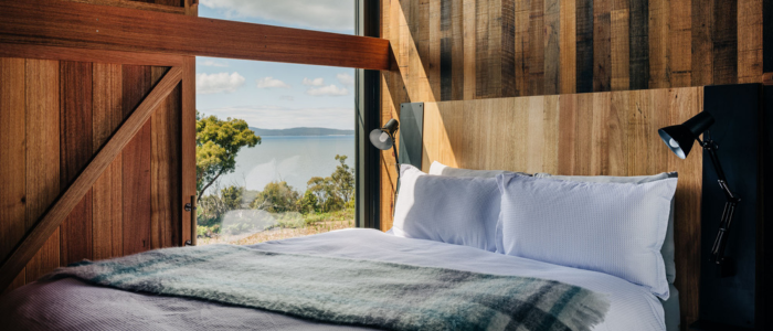 Cozy bedroom with wooden walls and ceiling, featuring a neatly made bed with white linens and a plaid blanket. Two adjustable reading lamps are mounted on the headboard. A large window offers a scenic view of a lake and lush greenery, blending rustic charm with natural beauty.