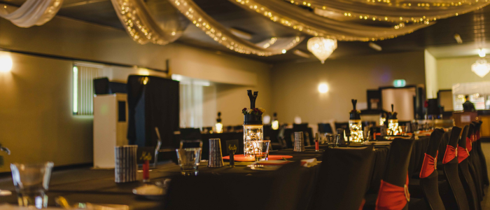 Decorated banquet hall with rows of tables covered in black cloths and glowing lantern centerpieces. Chairs are dressed in black covers with red sashes, and the ceiling features draped fabric and string lights, creating a warm, festive ambiance for a formal event.