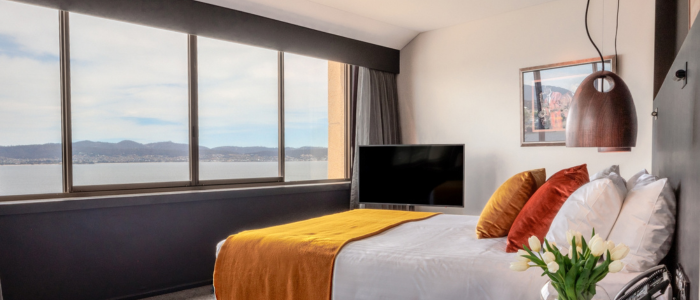 Stylish hotel bedroom with a large window overlooking water and hills. The room features a bed with white linens, orange accents, a wall-mounted TV, pendant light, bedside table with white tulips, and framed artwork.