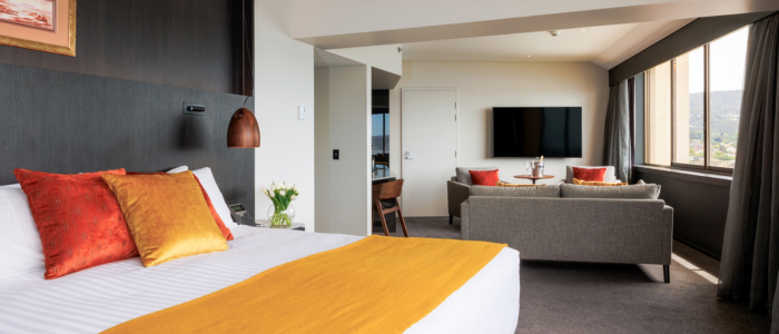 Modern hotel room with a large bed featuring white linens, an orange throw, and decorative pillows. The room includes a sitting area with a grey sofa, orange cushions, a round table with chairs, a wall-mounted TV, and large windows offering natural light and scenic views.