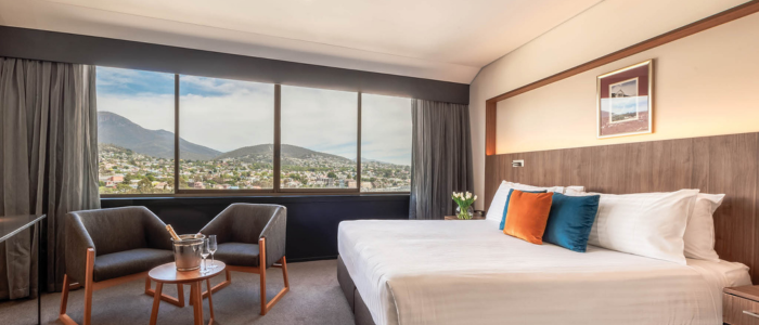 Bright hotel room with a large window overlooking a mountain and town view. The room includes a neatly made bed with white linens and colorful pillows, two armchairs, a round table with an ice bucket and glasses, and a framed picture above the bed.