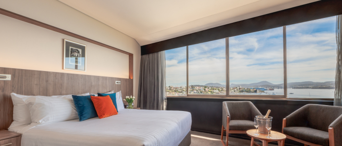 Bright hotel room with a large window overlooking a mountain and town view. The room includes a neatly made bed with white linens and colorful pillows, two armchairs, a round table with an ice bucket and glasses, and a framed picture above the bed.