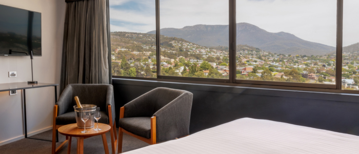 Hotel room with a large window showcasing a mountain and residential view. The room includes a bed with white linens, two gray armchairs around a small table with an ice bucket and wine glasses, and a wall-mounted flat-screen TV.