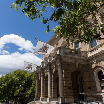 Hobart Town Hall 1 A historic sandstone building with classical architectural details, including tall columns and decorative stonework. Several flags reading “City of Hobart” fly from the façade. Tree branches frame the scene, and the sky is bright blue with scattered clouds.
