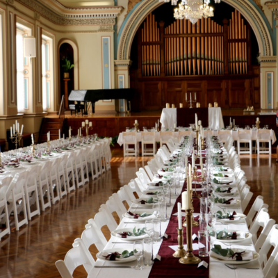 Hobart Town Hall 3 A grand reception hall arranged for a formal event, with two long rows of banquet tables covered in white tablecloths and burgundy runners. The tables are set with tall candles, glassware, and place settings. At the front of the room is a large, ornate pipe organ framed by an arched proscenium and a sparkling chandelier overhead.