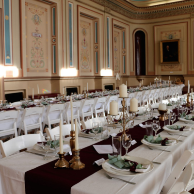 Hobart Town Hall 4 A long banquet table set for an event inside an ornate heritage hall. The table is dressed with white linens, a burgundy table runner, gold candlesticks, glassware, and neatly folded napkins with greenery. Rows of white chairs line the table, and the walls feature pastel-painted decorative panels and tall windows.