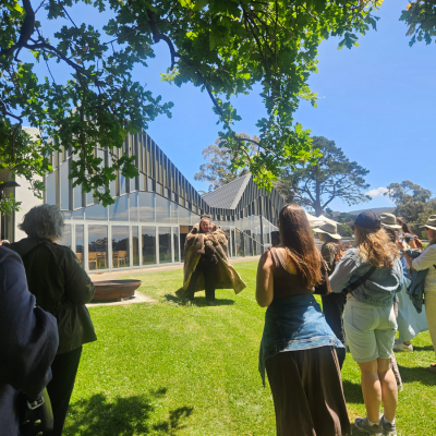 North America Famil Day 1.2 Group of people gathered outdoors on a grassy area near a modern glass building, watching a Welcome to Country Ceremony under a sunny blue sky with trees in the background.