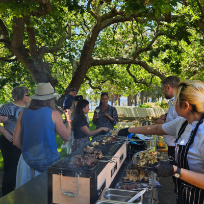 North America Famil Day 1.3 Group of people gathered around an outdoor barbecue under large leafy trees, with a chef grilling skewers on a stainless steel grill and tables set up in the background.