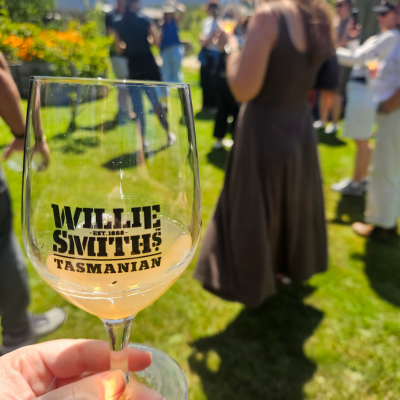 North America Famil Day 1.4 Close-up of a glass with ‘Willie Smith’s Tasmanian’ printed on it, held in the foreground with a light orange drink inside, while people stand on a grassy area in the background during an outdoor event.