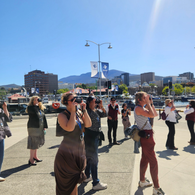 North America Famil Day 1.6 Group of people standing on a waterfront promenade taking photos, with boats docked in the harbor, city buildings, and mountains in the background under a clear blue sky.