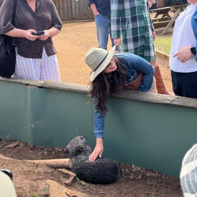 North America Famil Day 2.3 Person wearing a wide-brimmed hat and denim jacket leaning over a low enclosure to gently touch a wombat, with others standing nearby in an outdoor setting featuring wooden fencing and picnic tables.