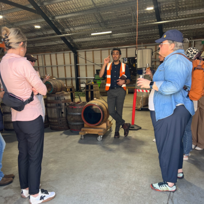 North America Famil Day 2.5 Group of people inside a warehouse-style building listening to a guide wearing a high-visibility vest, with wooden barrels and industrial equipment visible in the background.