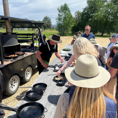 North America Famil Day 2.6 Outdoor cooking demonstration with a chef serving food from a large black barbecue trailer onto plates on a wooden table, surrounded by people wearing hats in a sunny open area with trees in the background.