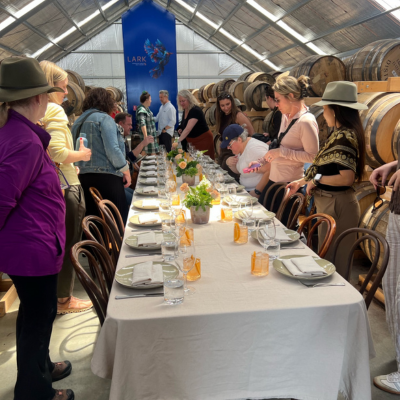 North America Famil Day 2.7 Group of people gathered around a long dining table set with white tablecloth, plates, glasses, and orange napkins inside a barrel room with wooden chairs and rows of large wooden barrels along the walls.
