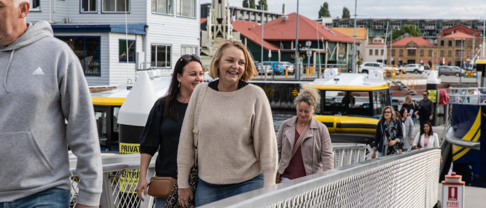 Pennicott Wilderness Journeys 1- The Bakery Creative Agency People walking along a metal gangway at a waterfront marina, with yellow and white boats docked nearby and buildings with red roofs in the background.