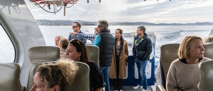 Pennicott Wilderness Journeys 2 - The Bakery Creative Agency Passengers seated inside a modern tour boat with large windows, while several people stand at the rear deck looking out over calm water toward distant hills under a partly cloudy sky.