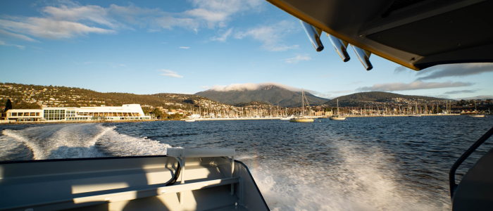 Pennicott Wilderness Journeys 3 - Beetle Black View from the back of a boat showing its wake trailing across calm blue water, with a marina full of sailboats and a coastal town in the background, and a mountain partially covered by clouds under a clear sky.
