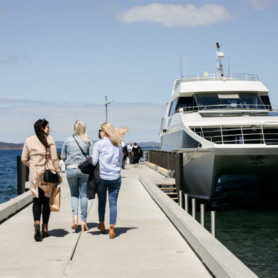 Peppermint Bay 1 Three people walking along a concrete pier toward a large white catamaran-style boat docked at the end, with calm blue water on both sides and a clear sky above.