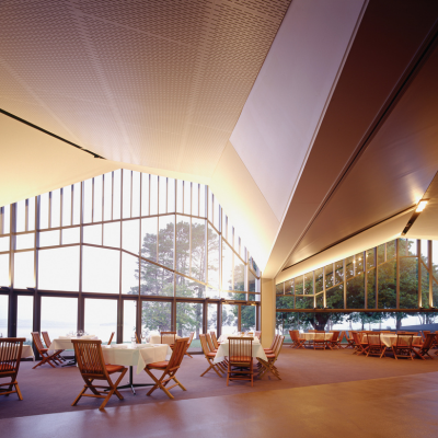 Peppermint Bay 2 Spacious modern dining area with high angled ceilings and large floor-to-ceiling windows overlooking trees and water, featuring neatly arranged tables with white tablecloths and wooden chairs under soft natural light.