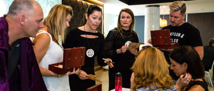 A small group of adults gathered closely around a table indoors, engaged in discussion during a workshop or activity. Several people hold open wooden boxes and cards, while one woman gestures as she speaks. Others listen attentively or read from the materials. The setting appears modern and professional, with warm lighting and a collaborative, problem-solving atmosphere.