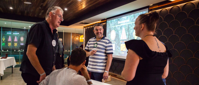 A small group of adults standing and seated around a table in a modern event space, engaged in conversation. One man smiles while holding a card, another speaks with animated body language, and a woman faces the group with her back partially to the camera. A large screen behind them displays colorful graphics, suggesting a facilitated workshop or group activity with an interactive, collaborative tone.