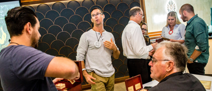 A group of adults interacting during a workshop in a modern indoor venue. A man at the center gestures while speaking, wearing a round badge, as others listen, handle small wooden boxes, or converse in pairs. The background features patterned wall panels and warm lighting, reinforcing a professional, collaborative event setting.