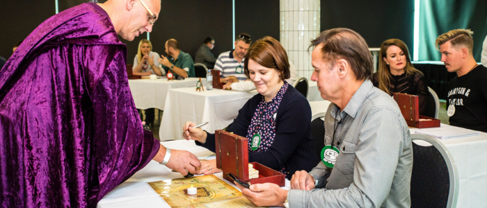 Two adults seated at a table participate in a hands-on workshop activity, examining a small wooden box and written materials. A facilitator wearing a purple cloak leans in to assist, holding an object over a board laid out on the table. Other participants work in the background, creating a focused, immersive atmosphere in a modern event space.