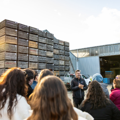 A guide speaks to a group outside Willie Smith’s cidery, with stacked wooden apple crates and the production shed behind him.