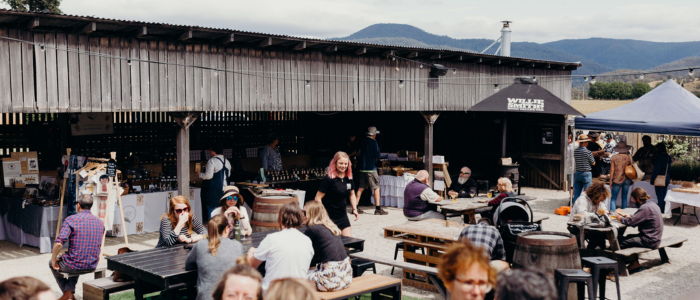 Guests gather at outdoor tables during an event at Willie Smith’s Apple Shed, with market stalls and mountain views in the background.