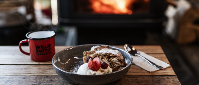 Apple pie served with ice cream and fresh berries beside a Willie Smith’s mug, with a wood fire glowing in the background.